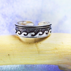 Può includere: Un anello regolabile color argento con un motivo a onde. L'anello presenta una fascia con una parte superiore strutturata e un motivo ripetuto di onde stilizzate. L'anello è esposto su una superficie di legno colorata chiara su uno sfondo blu e viola tenue.