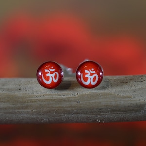 May include: A pair of silver stud earrings with red and white enamel. The earrings feature the Om symbol in white on a red background.