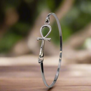 May include: A silver-colored bangle bracelet featuring an Ankh symbol. The Ankh, a symbol of life, is the focal point of the bracelet. The bracelet is displayed on a wooden surface with a blurred green background.