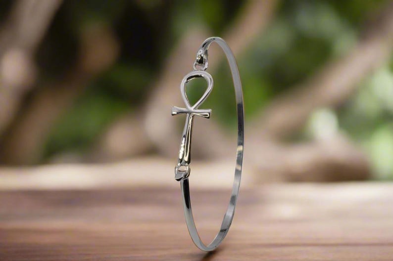 May include: A silver-colored bangle bracelet featuring an Ankh symbol. The Ankh, a symbol of life, is the focal point of the bracelet. The bracelet is displayed on a wooden surface with a blurred green background.