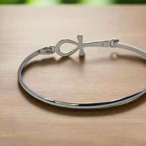 May include: A silver-colored bangle bracelet with an Ankh symbol at the center. The bracelet is circular and has a clasp closure. The Ankh symbol is a cross with a loop at the top. The bracelet is on a wooden surface.