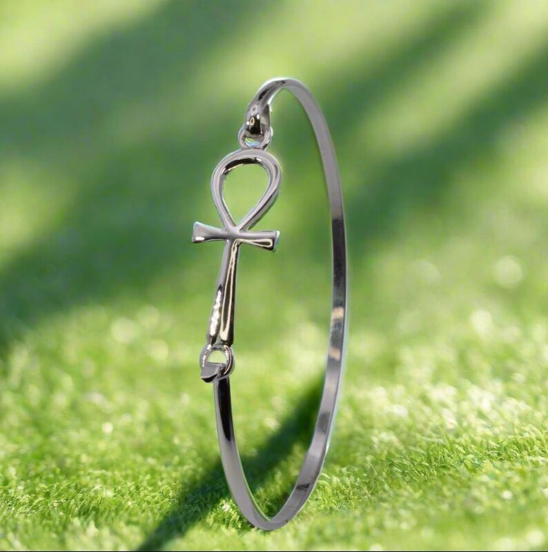May include: A silver-colored bangle bracelet featuring an Ankh symbol. The Ankh, a cross with a loop at the top, is a prominent design element. The bracelet is set against a green background, highlighting the jewelry's metallic sheen.