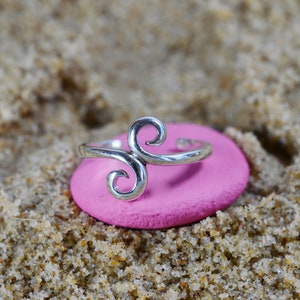 Puede incluir: Un anillo de dedo del pie de plata con un diseño de doble espiral, descansando sobre una superficie rosa.