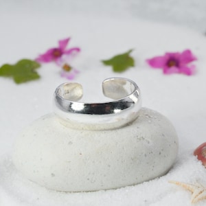 Puede incluir: Un brillante anillo de plata para el dedo del pie descansa sobre una piedra blanca texturizada. El anillo abierto refleja la luz. El fondo incluye flores rosas, hojas verdes y una estrella de mar, lo que sugiere un tema de playa. El anillo es sencillo y elegante.