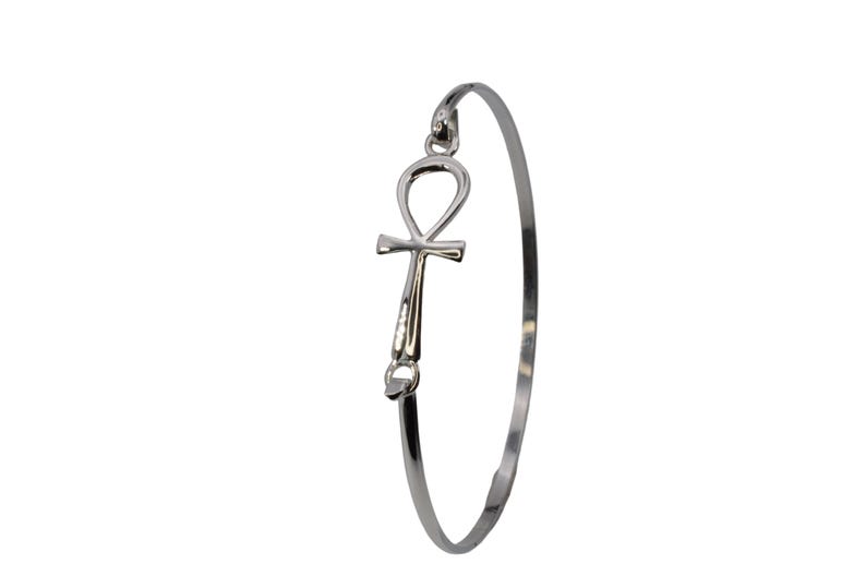 May include: A silver-colored bangle bracelet featuring an Ankh symbol. The Ankh, a symbol of life, is attached to the bracelet's band. The bracelet is a simple, elegant design, suitable for everyday wear.