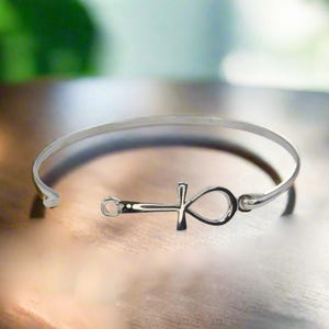 May include: A silver-colored bangle bracelet featuring an Ankh symbol. The open-ended cuff bracelet is made of a thin, shiny metal. The Ankh symbol is centered on the bracelet and is also silver-colored. The bracelet is displayed on a wooden surface.