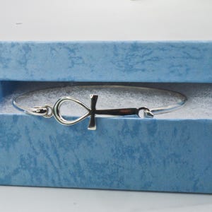 May include: A silver-colored ankh bracelet rests inside a light blue textured gift box. The ankh symbol is the focal point of the bracelet, with a loop at the top and a cross below. The box is lined with white, fluffy material.