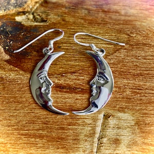 Puede incluir: Un par de pendientes de plata en forma de media luna con una cara esculpida en cada lado.