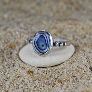 Peut inclure: Une bague en argent avec une incrustation de nacre d'ormeau bleue et verte. La bague est posée sur une pierre blanche et entourée de sable.