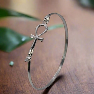 Puede incluir: Una pulsera de brazalete plateada con un símbolo Ankh. El Ankh, una cruz con un lazo, es el elemento central. La pulsera se muestra sobre una superficie de madera, con una hoja verde borrosa en el fondo.
