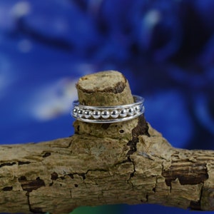 May include: A silver ring with a beaded band, sitting on a piece of weathered wood against a blue background.