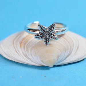 Puede incluir: Un anillo de plata con un diseño de estrella de mar, descansando sobre una concha blanca sobre un fondo azul.