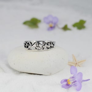 May include: A silver ring with an openwork design, resting on a white stone. The ring features a continuous pattern of swirling shapes. The background includes purple flowers and a starfish, suggesting a beach theme.