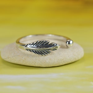 Puede incluir: Un anillo ajustable de plata con un diseño de pluma. El anillo está sobre una piedra blanca.