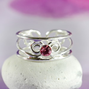 May include: A silver toe ring with a delicate, ornate design and a pink gemstone in the centre.