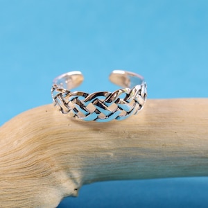 Puede incluir: Un anillo de dedo del pie de plata con un diseño trenzado. El anillo está sobre una superficie de madera marrón claro.