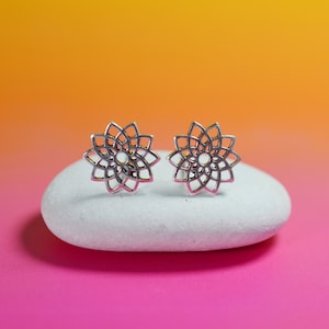 Op de afbeelding: Een paar zilveren oorbellen met een delicaat, ingewikkeld mandala-ontwerp. De oorbellen worden getoond op een witte steen tegen een roze en oranje achtergrond.