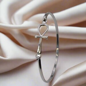 May include: A silver-colored bangle bracelet featuring an Ankh symbol. The bracelet is displayed against a backdrop of soft, cream-colored fabric. The Ankh symbol is a prominent focal point, adding a touch of ancient Egyptian design.