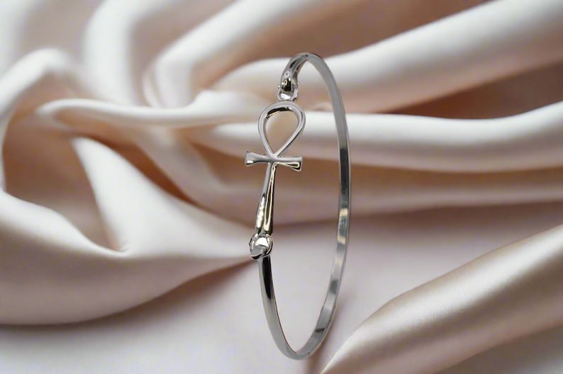May include: A silver-colored bangle bracelet featuring an Ankh symbol. The bracelet is displayed against a backdrop of soft, cream-colored fabric. The Ankh symbol is a prominent focal point, adding a touch of ancient Egyptian design.