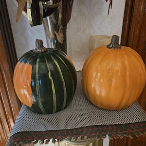 May include: Two decorative pumpkins with smooth, glossy surfaces. One pumpkin is primarily dark green with vertical orange stripes and a brown stem. The second pumpkin is solid orange with a brown stem. Both items feature rounded, ribbed shapes.