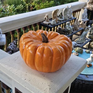 May include: A round, orange pumpkin decoration with a glossy finish and a dark brown, textured stem. The pumpkin features deep, vertical ridges and a smooth, rounded shape, capturing the classic appearance of a harvest-themed gourd.