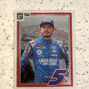 May include: A trading card featuring a NASCAR driver, Kyle Larson, wearing a blue racing suit with the number 5 on the front. The card is from the Optic series and is numbered 22.