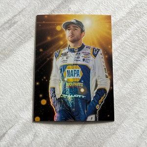 May include: A racing driver wearing a blue and yellow racing suit with the word "NAPA" on the front. The driver is standing in front of a gold and black background with a spotlight shining on him.