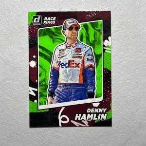 May include: A trading card featuring a green and black background with a photo of Denny Hamlin, a NASCAR driver, wearing a FedEx racing uniform. The card is from the Race Kings series.