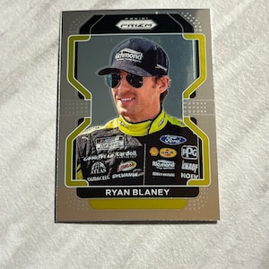 May include: A silver and yellow trading card featuring a photo of Ryan Blaney, a NASCAR driver, wearing a black cap with the word "Richmond" on it. The card has the word "PRIZM" in black letters on a silver background. The card also features the logos of various sponsors, including Goodyear, Cardell, Pennzoil, PPG, and Ford.
