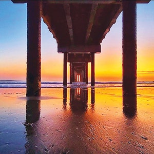 May include: A view of a wooden pier extending out over a sandy beach with the ocean in the background. The sky is a vibrant orange and blue, creating a beautiful sunset.
