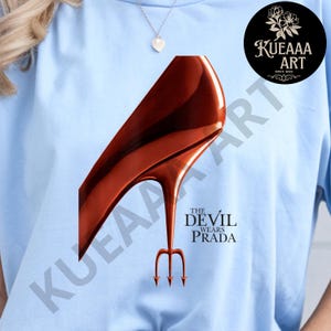 May include: Light blue t-shirt featuring a high-heeled shoe design in shades of brown and red, with a trident heel. The text "THE DEVIL WEARS PRADA" is printed below the shoe. A circular logo with text and floral design is in the upper right corner.