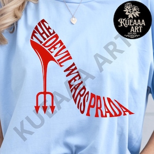 May include: Pale blue t-shirt with a red high-heeled shoe design. The shoe is formed by the words "THE DEVIL WEARS PRADA" and a trident heel. A small black circle with white text and floral design is in the upper right corner.