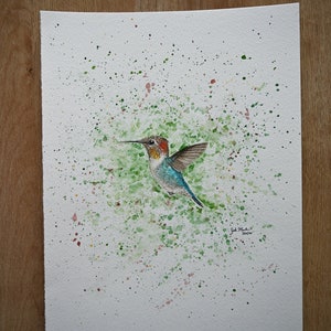 May include: Watercolor painting of a hummingbird in flight, surrounded by green and pink splatters on a white background. The artist's signature is in the bottom right corner.
