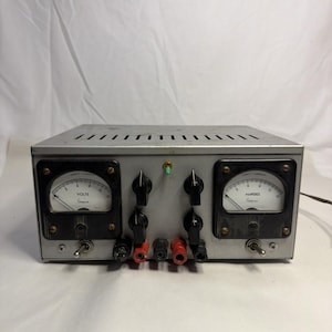 May include: A vintage silver-colored Simpson power supply unit. It features two analog meters, one for volts and one for amperes, with black knobs and terminals. The unit has a series of ventilation slots on top and a power switch.