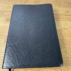 Puede incluir: Un libro negro cerrado con una cubierta texturizada. El libro tiene una cinta negra como marcador. La cubierta tiene una apariencia de cuero. El libro es rectangular y está sobre una superficie de madera.