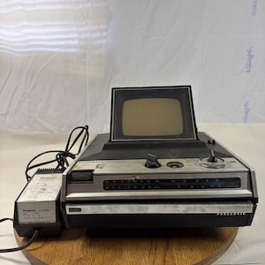 Puede incluir: Combinación de radio y televisión portátil Panasonic vintage. El dispositivo tiene una pantalla pequeña, un panel de control plateado y una base negra. Un adaptador de CA está conectado. La unidad está sobre una superficie de madera.