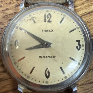 May include: Vintage Timex wristwatch with a cream-colored face, featuring black numerals at 12 and 3 o'clock, and black markers. The watch hands are silver. The word "TIMEX" is printed above the center, and "WATERPROOF" below. The watch has a silver band.