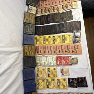 May include: A collection of vintage guitar and string instrument strings. The packaging includes boxes and individual string packets in various colors, including black, yellow, and brown. The brands include Black Diamond, Mapes, and Resnah.