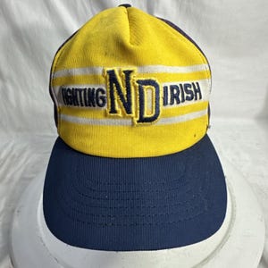 Puede incluir: Gorra de béisbol amarilla y azul marino con el texto "FIGHTING IRISH ND" en azul marino y blanco. La gorra tiene una visera curva y una corona amarilla con rayas blancas.