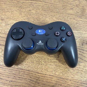 May include: Black Logitech PlayStation controller with blue accents. Features a directional pad, analog sticks, and action buttons. The controller has the PlayStation logo and the Logitech logo.