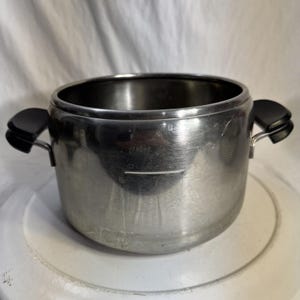 Puede incluir: Una olla de cocina de acero inoxidable con dos asas negras. La olla tiene forma cilíndrica y una abertura ancha. Una línea de medición es visible en el lateral. La olla está sobre una superficie blanca.