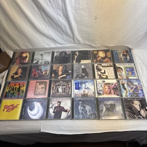 Puede incluir: Una colección de CD de música, con varios artistas y portadas de álbumes. Los CD están en estuches de plástico transparente, con ilustraciones y texto coloridos. La colección incluye una variedad de géneros, incluyendo pop, rock y música dance.