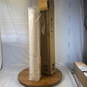May include: A tall, cylindrical roll of white paper wrapped in clear plastic, standing vertically next to its tall, rectangular cardboard shipping box. The box is tan with black HP logos and various shipping icons printed on the front side.