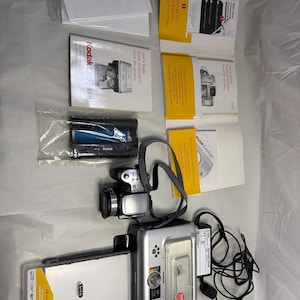 Puede incluir: Una cámara digital y una impresora Kodak EasyShare plateadas con varios accesorios. Incluye guías del usuario, un CD, un cargador de batería y una correa para la cámara. La impresora tiene una cubierta transparente y una etiqueta amarilla.