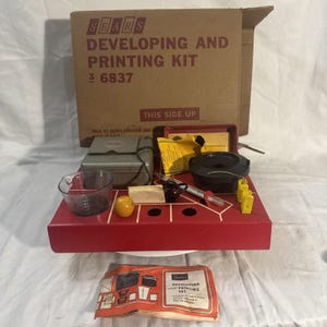 May include: A vintage Sears Developing and Printing Kit, featuring a cardboard box with text, a red tray with various tools, a clear measuring cup, and a small orange ball. The kit includes a darkroom enlarger and other accessories.
