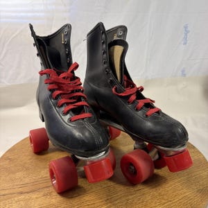 Könnte beinhalten: Ein Paar Vintage-Rollschuhe. Die schwarzen Rollschuhe haben rote Schnürsenkel und rote Rollen. Die hoch geschnittenen Stiefel weisen sichtbare Nähte und Metallösen für die Schnürsenkel auf. Die Rollen haben ein klassisches, abgerundetes Design.