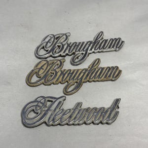 May include: Three vintage car emblems. Two are labeled "Brougham" in a silver script font, and one is labeled "Fleetwood" in a similar font. The emblems are slightly tarnished, showing signs of age and use. The background is a light, neutral color.