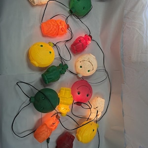 May include: A string of vintage Christmas lights featuring various colorful plastic figures. The lights include green, orange, yellow, red, and white shapes, connected by a black wire. The lights are lit, casting a warm glow.
