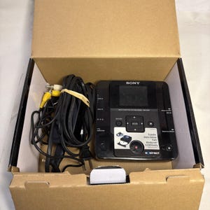May include: A black Sony multi-function DVD recorder, model VRD-MC6, sits inside a cardboard box. Cables and connectors are visible, including yellow and white RCA cables. The device has a screen and several buttons.