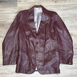Puede incluir: Una blazer de cuero burdeos con cierre clásico de tres botones y solapas con muescas. La chaqueta presenta dos bolsillos delanteros y un forro de color claro. La blazer es de estilo vintage.